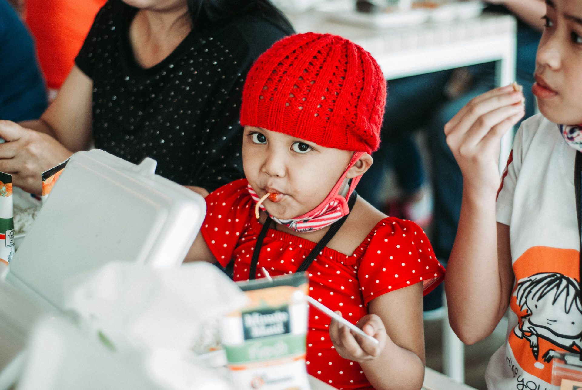 child wearing red and white dress eating while sitting near two person